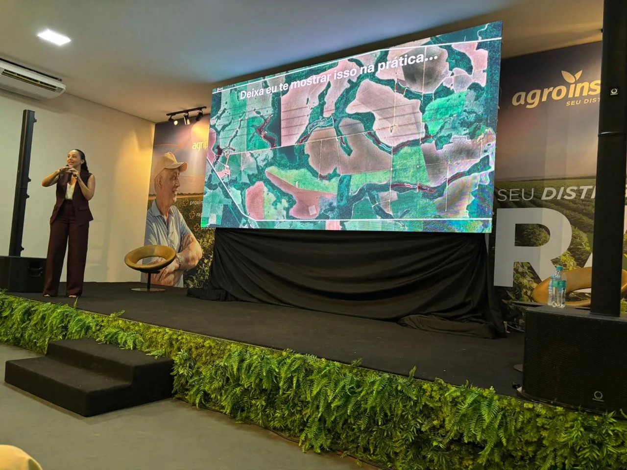 Soraia no palco ao lado de um telão com imagem de satélite de propriedades rurais